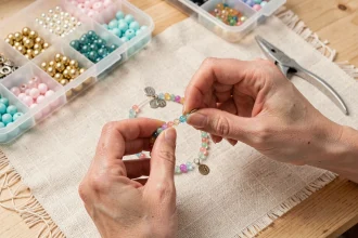 Mains de femme enfilant des perles colorées et des breloques dorées sur un élastique pour créer un bracelet personnalisé sur un établi en bois clair