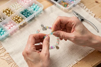 Mains de femme enfilant des perles colorées et des breloques dorées sur un élastique pour créer un bracelet personnalisé sur un établi en bois clair